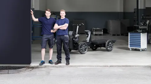 Two service technicians are standing in front of a workshop housing an eCargo bike