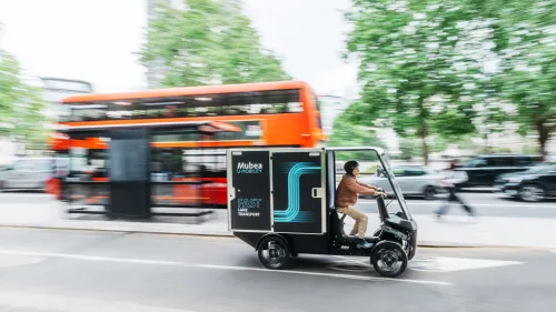 Electric cargo vehicle in motion on a city street, passing a bus stop and double-decker bus