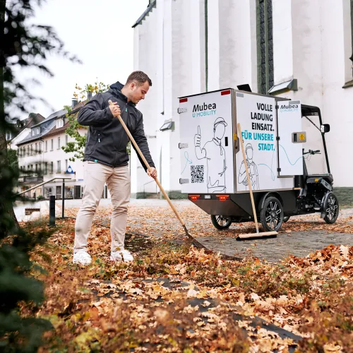A young worker sweeps autumn leaves in front of an eCargobike.