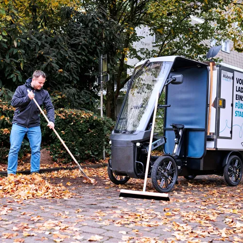 Young worker sweeping leaves, in the background is his eCargobike with other street cleaning tools.
