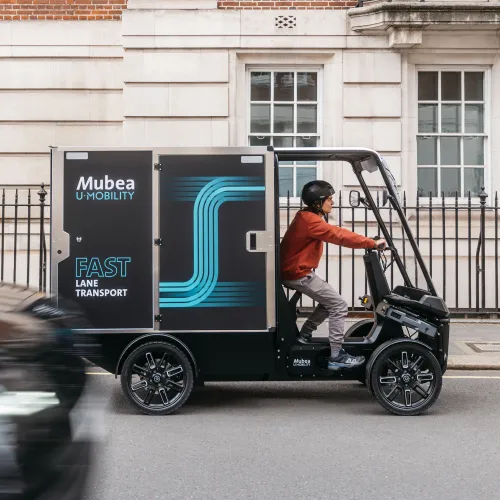 Electric cargo vehicle travels along a city street, passing historic buildings and traffic.