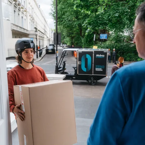 Delivery worker hands over a package at a doorstep beside an electric cargo vehicle.