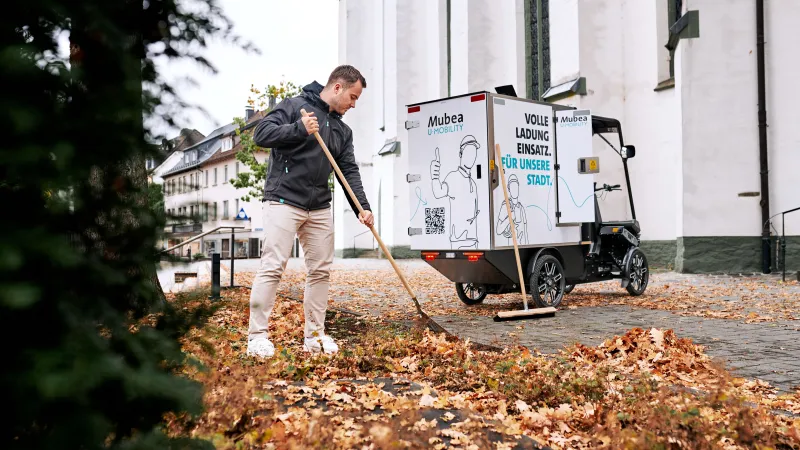 A young worker sweeps autumn leaves in front of an eCargobike.