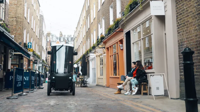 Electric cargo vehicle drives through a narrow cobbled street lined with cafés and shops.